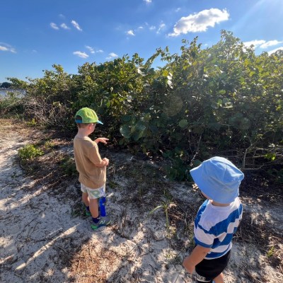 Two children in hats exploring a sandy beach path with greenery and water nearby.