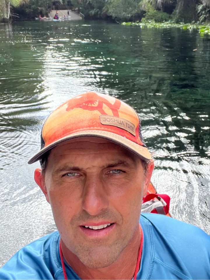 Man in a hat and blue shirt near a body of water with greenery.