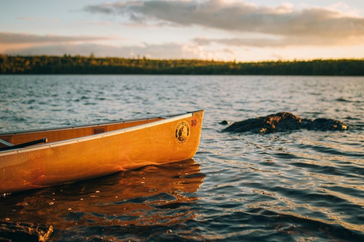 A canoe on a lake at sunset with a rocky shore and trees in the background.