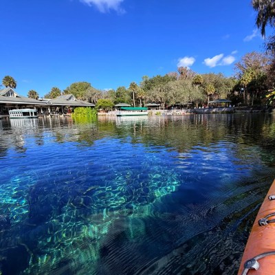 Clear water with a yellow kayak, buildings and trees in the background under a blue sky.