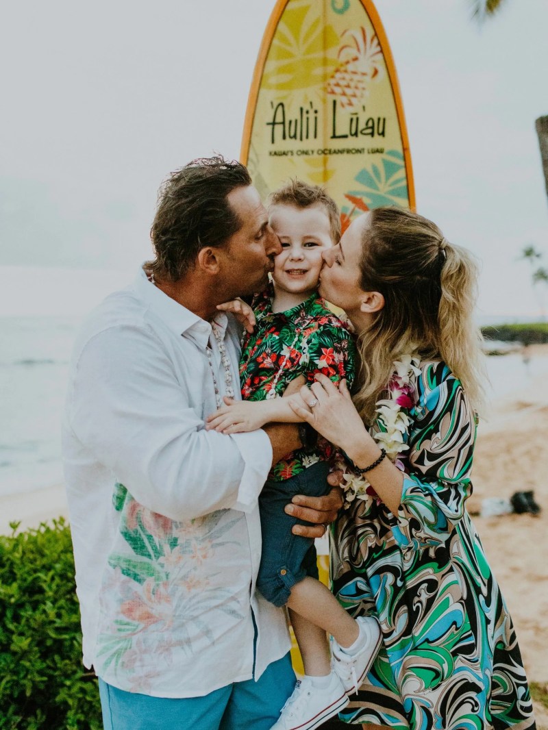 Family with surfboard, parents kissing child on the beach.