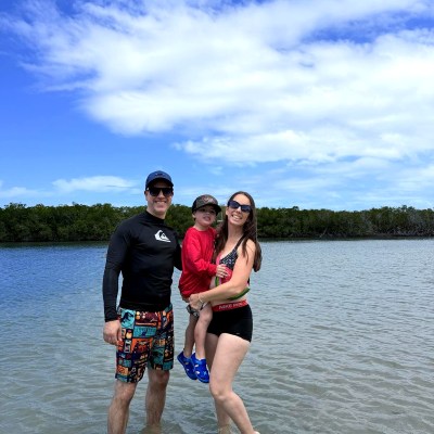 Smiling family standing in shallow water under a blue sky.