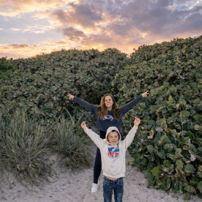 Woman and child on sandy path with lush greenery and colorful sunset sky.