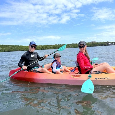 Family of three kayaking on calm water under a blue sky.