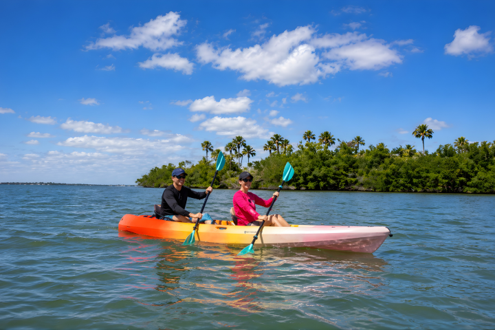 Two people kayaking on a calm lake under a blue sky with scattered clouds and trees in the background.