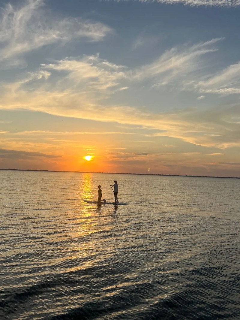 Two people stand-up paddleboarding on calm water at sunset.